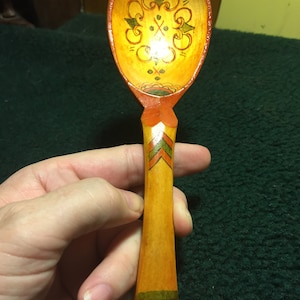 May include: Hand-painted wooden spoon with a yellow bowl and handle. The bowl features a decorative floral design in green and orange. The handle has orange, green, and yellow painted details. A unique kitchen utensil.
