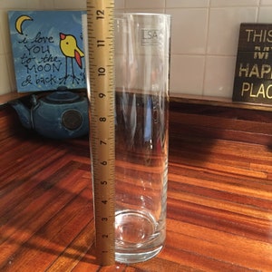 May include: A clear glass cylinder vase, 12 inches tall, with a round base. The vase is sitting on a wooden surface.