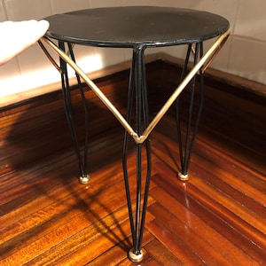 May include: A small, round side table with a black top and black metal legs. The legs are accented with gold-colored metal and have gold-colored feet. The table has a mid-century modern design.