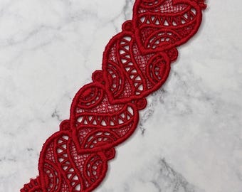 May include: A red, embroidered bookmark featuring a series of connected heart shapes. The bookmark is made of delicate lace-like material with intricate patterns and a red ribbon for hanging. The design is set against a marble background.