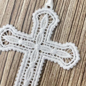 May include: A white, intricately designed lace cross with a satin ribbon for hanging. The cross features detailed openwork patterns and a delicate, textured appearance. The background is a light brown wood grain.