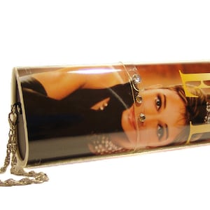 May include: A cylindrical clutch bag featuring a glossy photographic print of a person on the exterior. The bag includes a metallic chain strap and a clear plastic band adorned with small circular embellishments across the center surface.