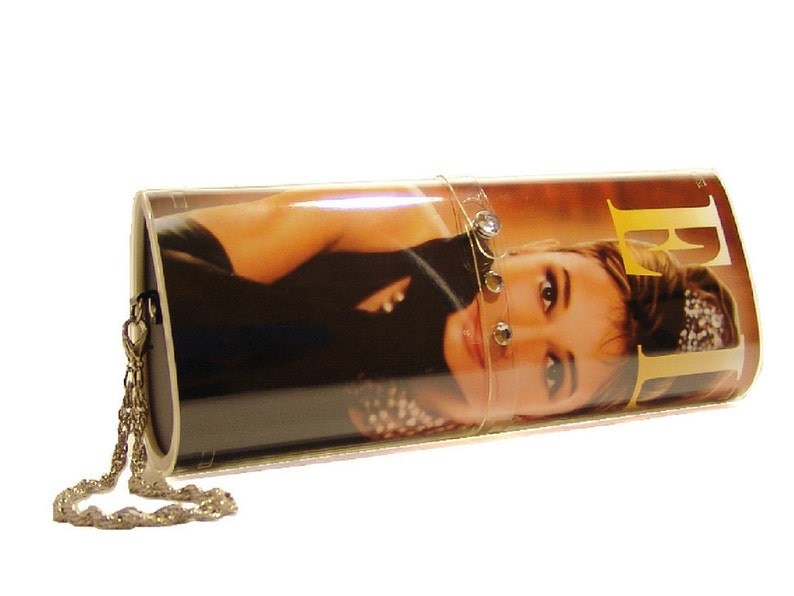 May include: A cylindrical clutch bag featuring a glossy photographic print of a person on the exterior. The bag includes a metallic chain strap and a clear plastic band adorned with small circular embellishments across the center surface.
