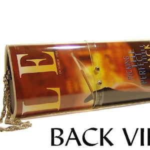 May include: A cylindrical clutch bag featuring a glossy cover of an October 1961 Elle magazine with the text Picking The Perfect Little Black Dress. The bag has a clear plastic strap with two metal studs and a thin gold-toned metal chain attached.