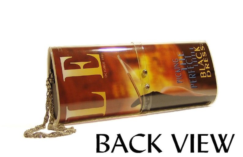 May include: A cylindrical clutch bag featuring a glossy cover of an October 1961 Elle magazine with the text Picking The Perfect Little Black Dress. The bag has a clear plastic strap with two metal studs and a thin gold-toned metal chain attached.
