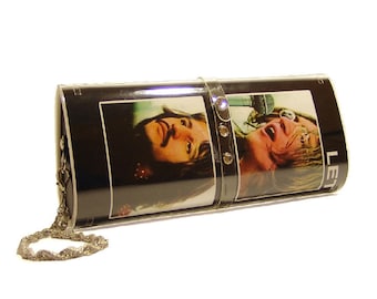 May include: A rectangular black clutch bag featuring two photographic portraits of individuals with long hair. A clear plastic strap with three small circular embellishments secures the center. A silver-toned metal chain is attached to the side.