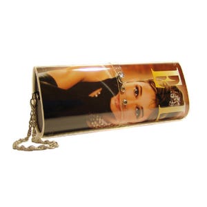 May include: A cylindrical clutch bag featuring a glossy print of a woman on the cover of Elle magazine. The bag has a gold-toned metal chain strap and a clear plastic band across the centre adorned with three small, clear, round decorative stones.