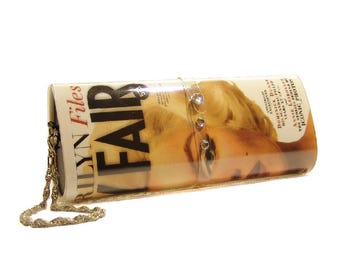 May include: A rectangular clutch bag featuring a glossy print of a magazine cover with the text "VANITY FAIR" and "Marilyn Files". The bag has a metallic gold chain strap and a clear plastic strap across the center adorned with three small rhinestones.