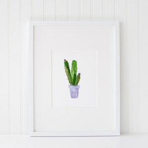 May include: A watercolor painting of a green cactus plant in a purple pot with pink and purple flowers. The painting is framed in a white frame.