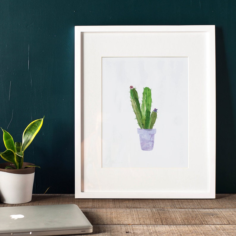 May include: A watercolor painting of a cactus in a purple pot with pink and purple flowers. The painting is framed in a white frame and is hanging on a green wall.