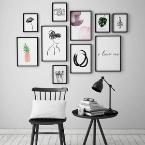 Op de afbeelding: Een zwarte stoel met een wit kussen staat voor een witte muur met zwart ingelijste kunstwerken. De kunstwerken omvatten een camera, een aquarel, een monstera-blad, een vrouwensilhouet, een cirkel en een print die "i love us" zegt. Een zwarte bijzettafel met een lamp en boeken staat rechts van de stoel.