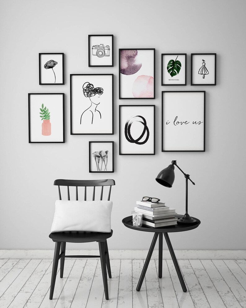 Op de afbeelding: Een zwarte stoel met een wit kussen staat voor een witte muur met zwart ingelijste kunstwerken. De kunstwerken omvatten een camera, een aquarel, een monstera-blad, een vrouwensilhouet, een cirkel en een print die "i love us" zegt. Een zwarte bijzettafel met een lamp en boeken staat rechts van de stoel.