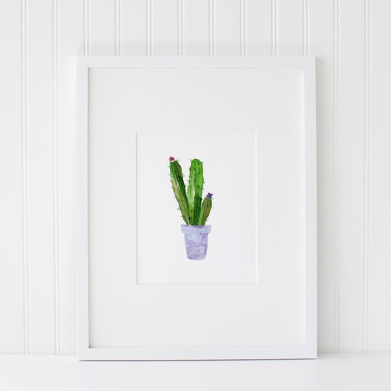 May include: A watercolor painting of a green cactus plant in a purple pot with pink and purple flowers. The painting is framed in a white frame.