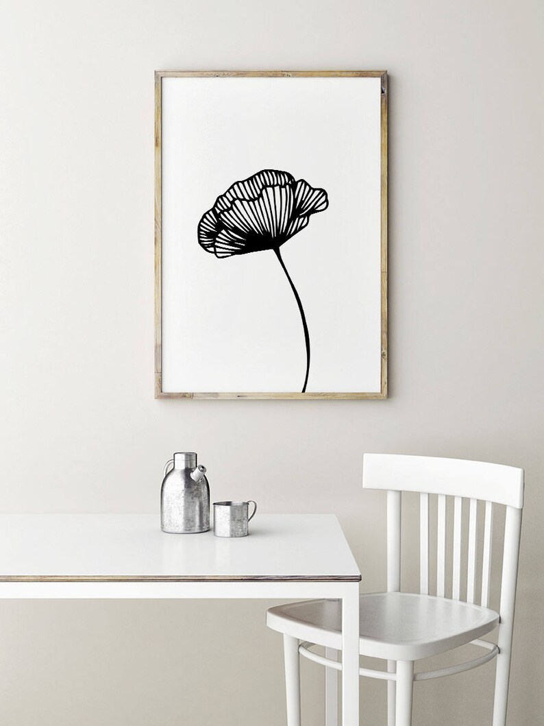 Op de afbeelding: Een zwart-witte lijntekening van een klaproos in een houten lijst die aan een witte muur hangt boven een witte tafel met een zilveren kan en mok.