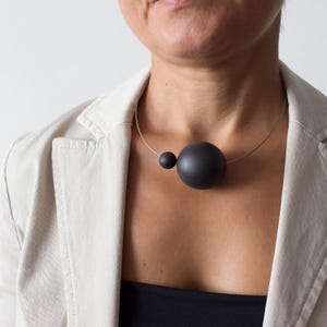 Könnte beinhalten: Eine schwarze Choker-Kette mit zwei großen schwarzen runden Perlen. Die Kette wird von einer Person mit einem hellbeigen Blazer getragen.
