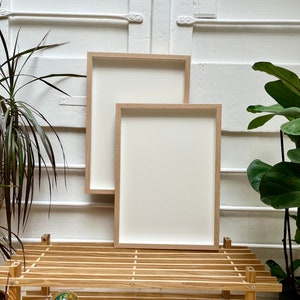 Op de afbeelding: Twee lege, natuurlijke houten fotolijsten met witte passe-partouts staan op een houten plank. De lijsten zijn rechthoekig en hebben een eenvoudig, modern ontwerp.