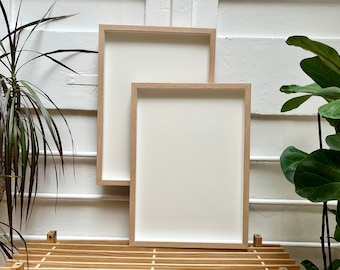 Puede incluir: Dos marcos de fotos vacíos de madera natural con paspartús blancos están apilados en un estante de madera. Los marcos son rectangulares y tienen un diseño simple y moderno.