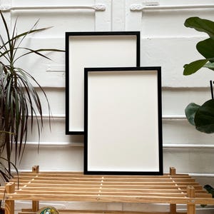 May include: Two black picture frames of different sizes are stacked on top of each other on a wooden shelf. The frames are empty and have a white background.