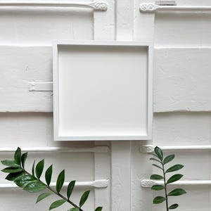 May include: A white picture frame with a white mount is hanging on a white wall. The frame is empty and ready for a photo or artwork.