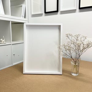 May include: A large, white picture frame with a simple, modern design. The frame is rectangular and sits on a brown surface. A small glass vase with delicate white flowers is to the right of the frame. Several framed pictures are on the wall.