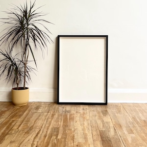 May include: A black picture frame with a white mount is standing on a wooden floor. A potted plant is in the background.