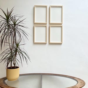 May include: Four rectangular wooden picture frames with white interiors are mounted on a white wall. A tall potted plant with long, thin green leaves sits to the left. A round wooden table with a glass top is in the foreground.