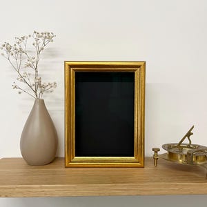 以下が含まれることがあります： 黒い内側の金色の額縁が木製の棚に置かれています。白い花が入った花瓶と真鍮の日時計も棚にあります。フレームは装飾的なデザインです。