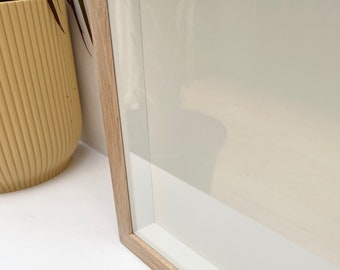 Puede incluir: Primer plano de un marco de imagen de madera clara con un passe-partout blanco y un frente de vidrio transparente. El marco está sobre una superficie blanca.