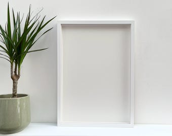 以下が含まれることがあります： 白いマットが付いた白い写真フレームが、左側に緑色の鉢植えの植物がある白い棚の上に置かれています。
