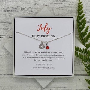May include: A silver necklace with a small charm that says "A" and a red ruby gemstone. The necklace comes in a white box with a card that says "July Ruby Birthstone" and describes the meaning of the ruby.