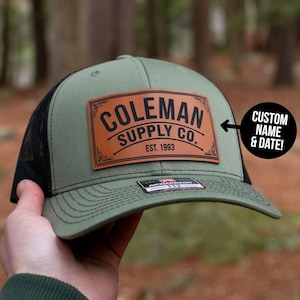 Puede incluir: Gorra de camionero verde oliva y negra con un parche de cuero marrón. El parche presenta el texto "COLEMAN SUPPLY CO. EST. 1993" en letras negras. La gorra tiene una parte trasera de malla y una visera curva. También se ve el texto "CUSTOM NAME & DATE!".