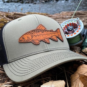 May include: A green and black trucker hat with a brown leather patch of a trout fish. The hat is sitting on a bed of brown leaves and twigs.
