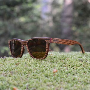 May include: Wooden sunglasses with brown and red frames and brown lenses. The sunglasses are resting on a bed of green moss.