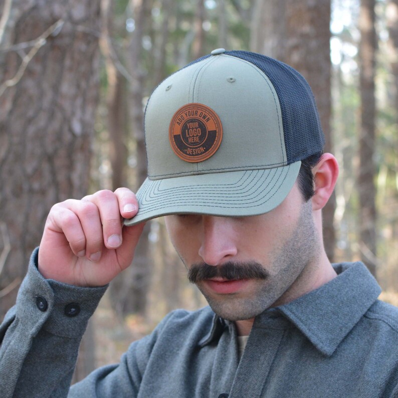 May include: A green and black trucker hat with a brown leather patch that says 'Add Your Own Design' and 'Your Logo Here'.