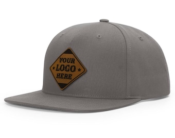 May include: A gray flat brimmed baseball cap with a brown leather patch that says "YOUR LOGO HERE" in black lettering.