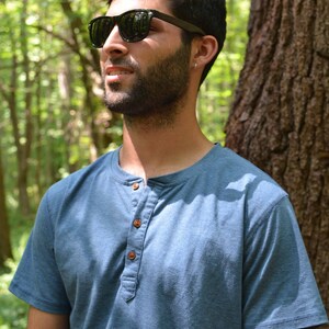 May include: A man wearing a blue short-sleeved shirt with a button-up collar and dark sunglasses. He has a beard and is standing in a forest.