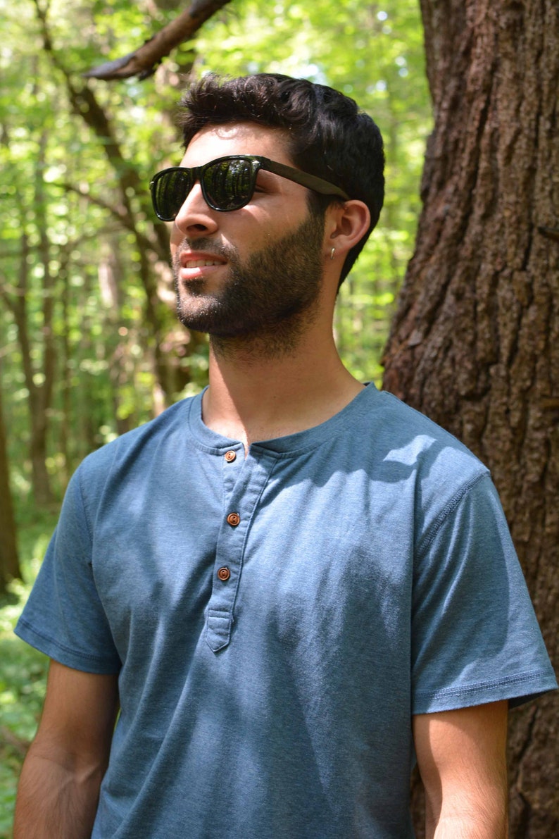 May include: A man wearing a blue short-sleeved shirt with a button-up collar and dark sunglasses. He has a beard and is standing in a forest.
