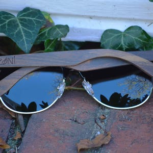 Puede incluir: Un par de gafas de sol con montura de madera oscura y lentes negras. Las gafas est&aacute;n dobladas por la mitad y el nombre de la marca "ZONI" es visible en la montura.
