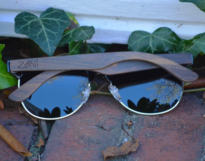 Puede incluir: Un par de gafas de sol con montura de madera oscura y lentes negras. Las gafas est&aacute;n dobladas por la mitad y el nombre de la marca "ZONI" es visible en la montura.