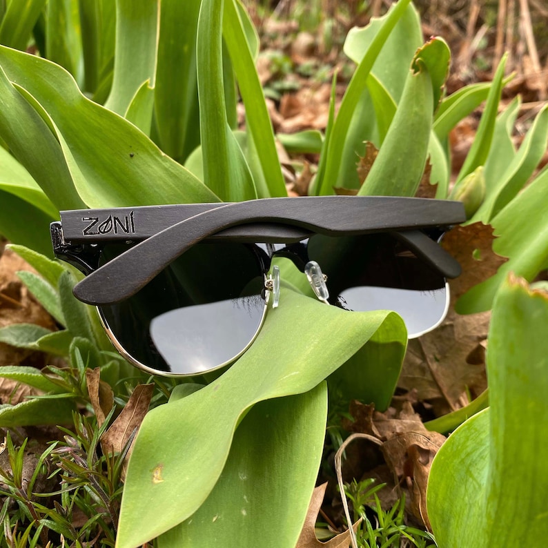 Puede incluir: Gafas de sol de madera negra con un logotipo ZONI en el lateral. Las gafas de sol est&aacute;n apoyadas en hojas verdes.