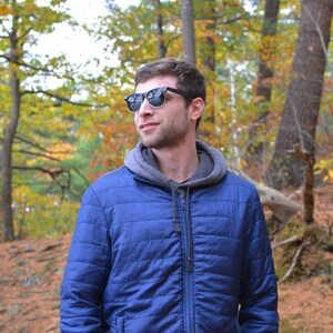 Puede incluir: Un hombre con una chaqueta azul con capucha y gafas de sol est&aacute; de pie en un bosque. Los &aacute;rboles en el fondo tienen follaje de oto&ntilde;o.
