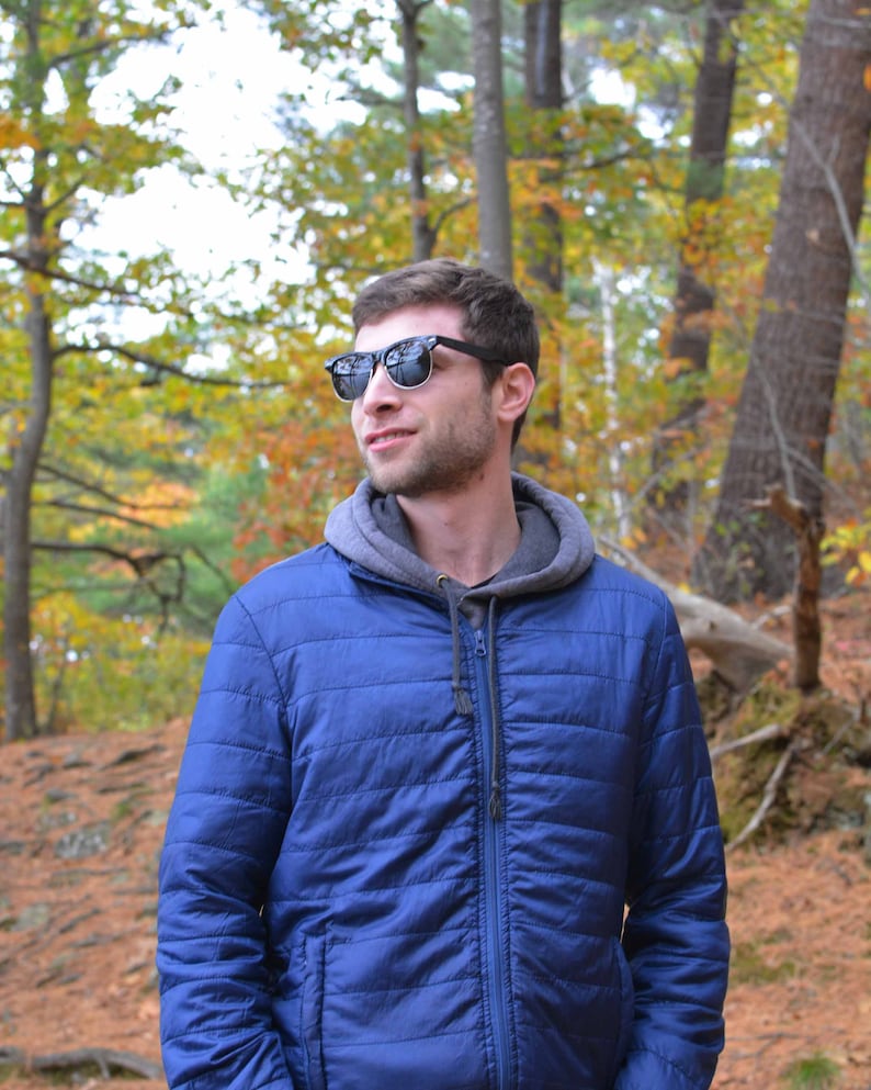 Puede incluir: Un hombre con una chaqueta azul con capucha y gafas de sol est&aacute; de pie en un bosque. Los &aacute;rboles en el fondo tienen follaje de oto&ntilde;o.