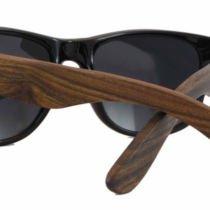 May include: Black sunglasses with dark lenses and wooden arms. The arms are made of dark brown wood with a natural grain pattern. The brand name "Zani" is engraved on the left arm.