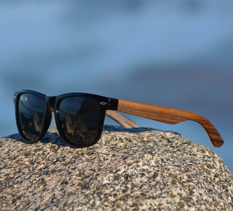 May include: Black sunglasses with wooden arms. The sunglasses have a dark lens and the arms are made of natural wood. The brand name "ZONI" is engraved on the right arm.