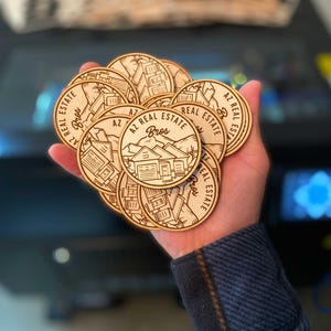 May include: A hand holding a stack of round wooden coasters with a laser-engraved design. The coasters feature the text "AZ Real Estate Bros" and a stylized image of a house.