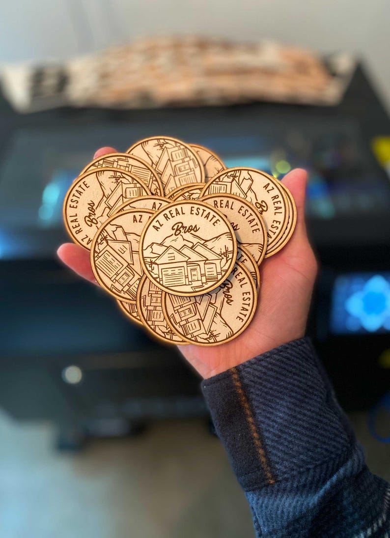 May include: A hand holding a stack of round wooden coasters with a laser-engraved design. The coasters feature the text "AZ Real Estate Bros" and a stylized image of a house.
