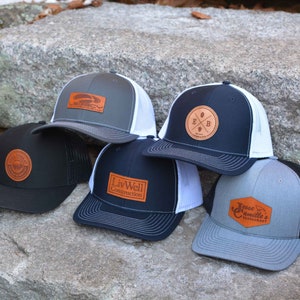 May include: Five different trucker hats with leather patches. The patches feature various logos and text, including "Liv Well Construction", "E & B", "Jesse Camille's Restaurant", and a logo with a plane.