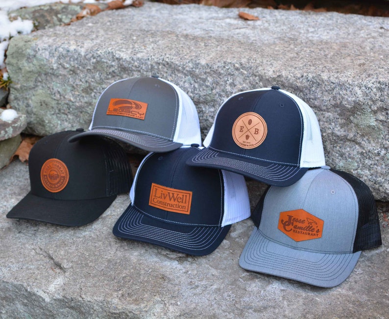 May include: Five different trucker hats with leather patches. The patches feature various logos and text, including "Liv Well Construction", "E & B", "Jesse Camille's Restaurant", and a logo with a plane.