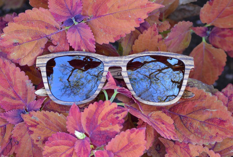 May include: Wooden sunglasses with brown and black striped frames and blue lenses. The sunglasses are lying on a bed of red and purple leaves.