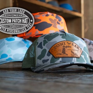 Może przedstawiać: Trzy truckerki w kamuflażu. Środkowa czapka ma brązową skórzaną naszywkę z tekstem "ZONI WEAR Outfitters EST. 2015 NEW HAMPSHIRE".